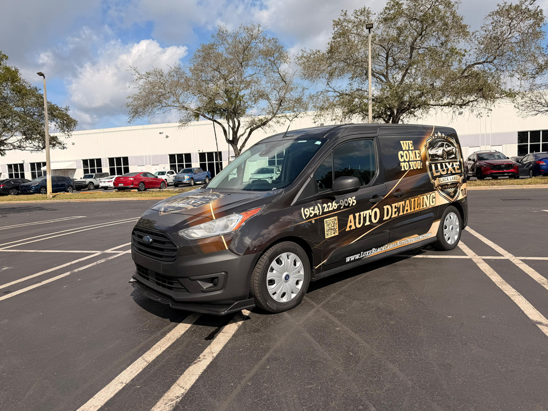 Brown/black Luxe Auto Detailing van with 'AUTO DETAILING' in gold, parked in a lot; shows phone number and website on the side.