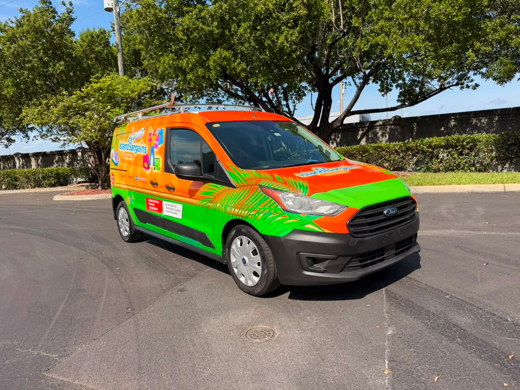 Bright orange and green promotional van with tropical graphics parked in a paved lot, trees in the background.