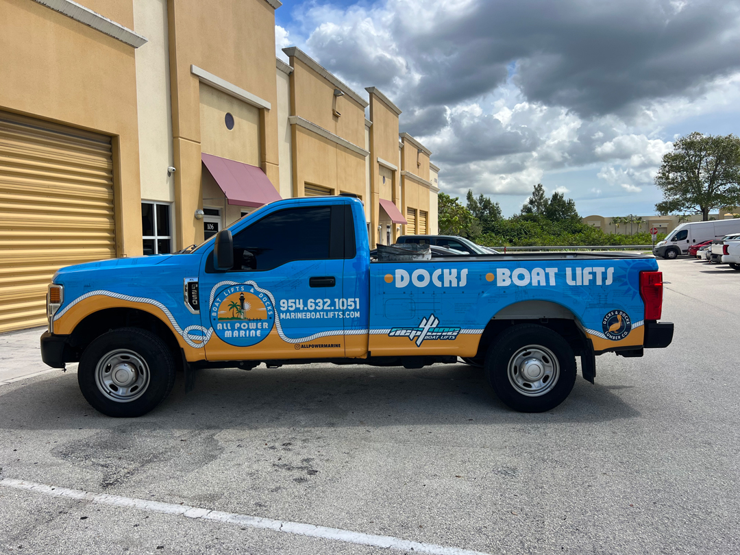 Blue and tan service pickup truck with 'Docks Boat Lifts' signage parked in a storage complex; features contact info and boat-lift logos on the side panel.