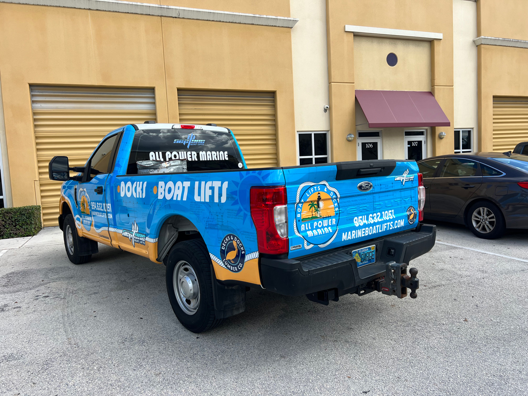 Blue promotional pickup wrapped with 'Dock & Boat Lifts' branding and marine service logos, parked by a storage building.