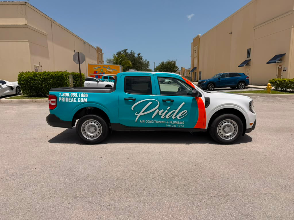 Teal Pride air conditioning and plumbing service pickup truck with large script logo and contact info parked on a sunny street in front of beige buildings.