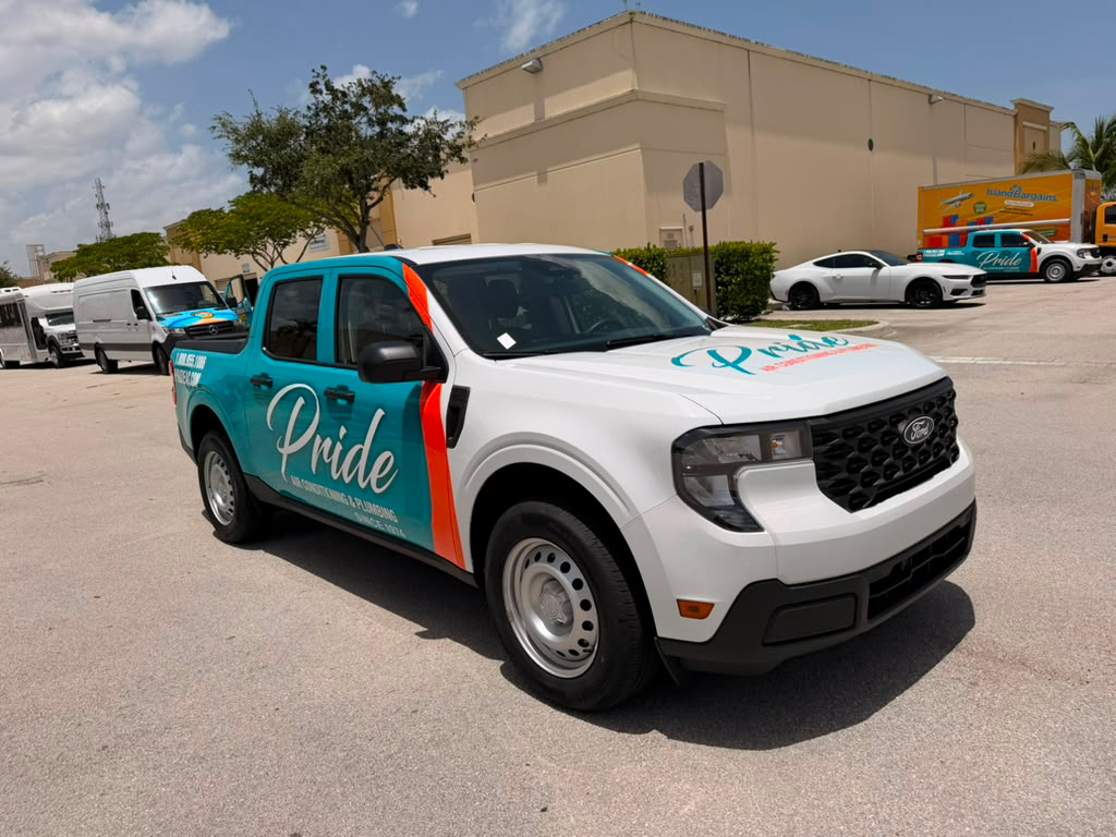 White Ford cargo van with teal Pride branding parked in an outdoor lot amid other vehicles; moving company vehicle in use.