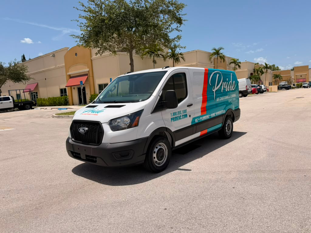 White Pride van parked in a sunny lot beside a beige shopping plaza with palm trees in the background, side of van shows Pride branding and contact info.