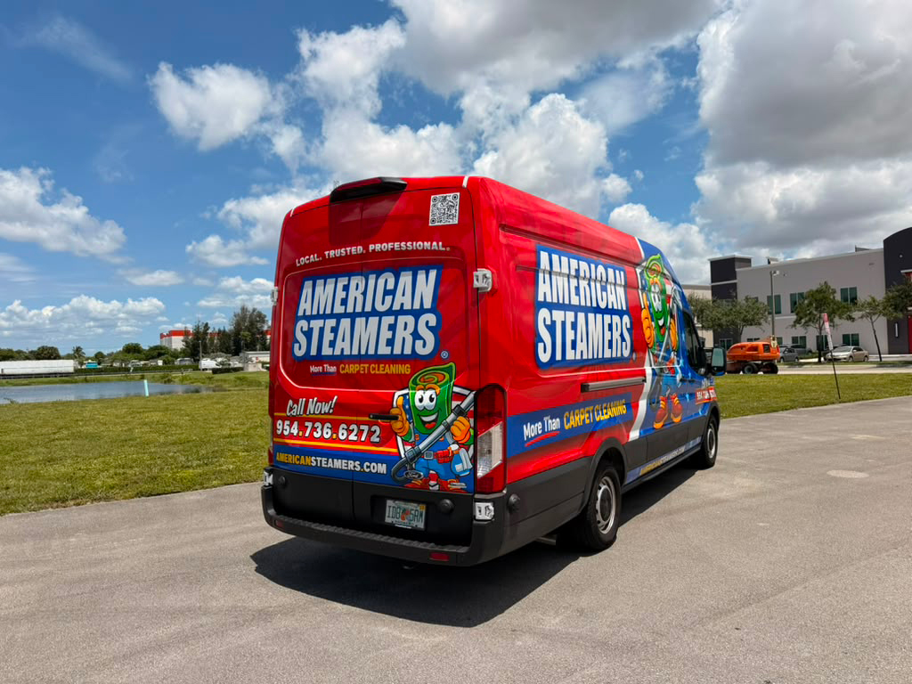 Red carpet-cleaning van with 'American Steamers' branding parked in a lot on a sunny day, featuring a cartoon mascot and contact info.